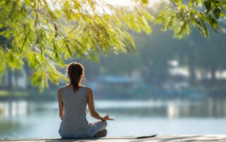 Rückenbild einer Frau, die für verbessertes Stressmanagement für Führungskräfte vor einem See in der Natur meditiert