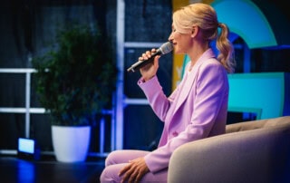 Kerstin Hardt in einem fliederfarbenen Blazer sitzend auf der Bühne von Greator erklärt die Symptome Stress