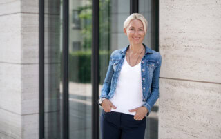 Kerstin Hardt, Spezialistin für Ernährung zum Abnehmen, steht in einer Jeansjacke lächelnd vor einer Glasfassade.